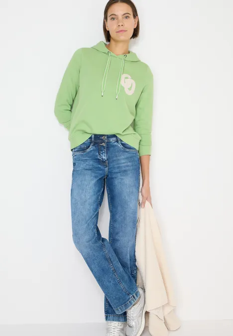 Person trägt hellgrünen Hoodie, blaue Jeans und hält einen beigen Mantel.